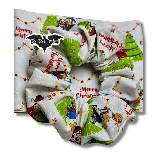White fabric scrunchie with green Christmas trees, candy-cane details, and twinkle-light garland over “Merry Christmas” text; handmade holiday hair tie by Boo Boo’s Creations.
